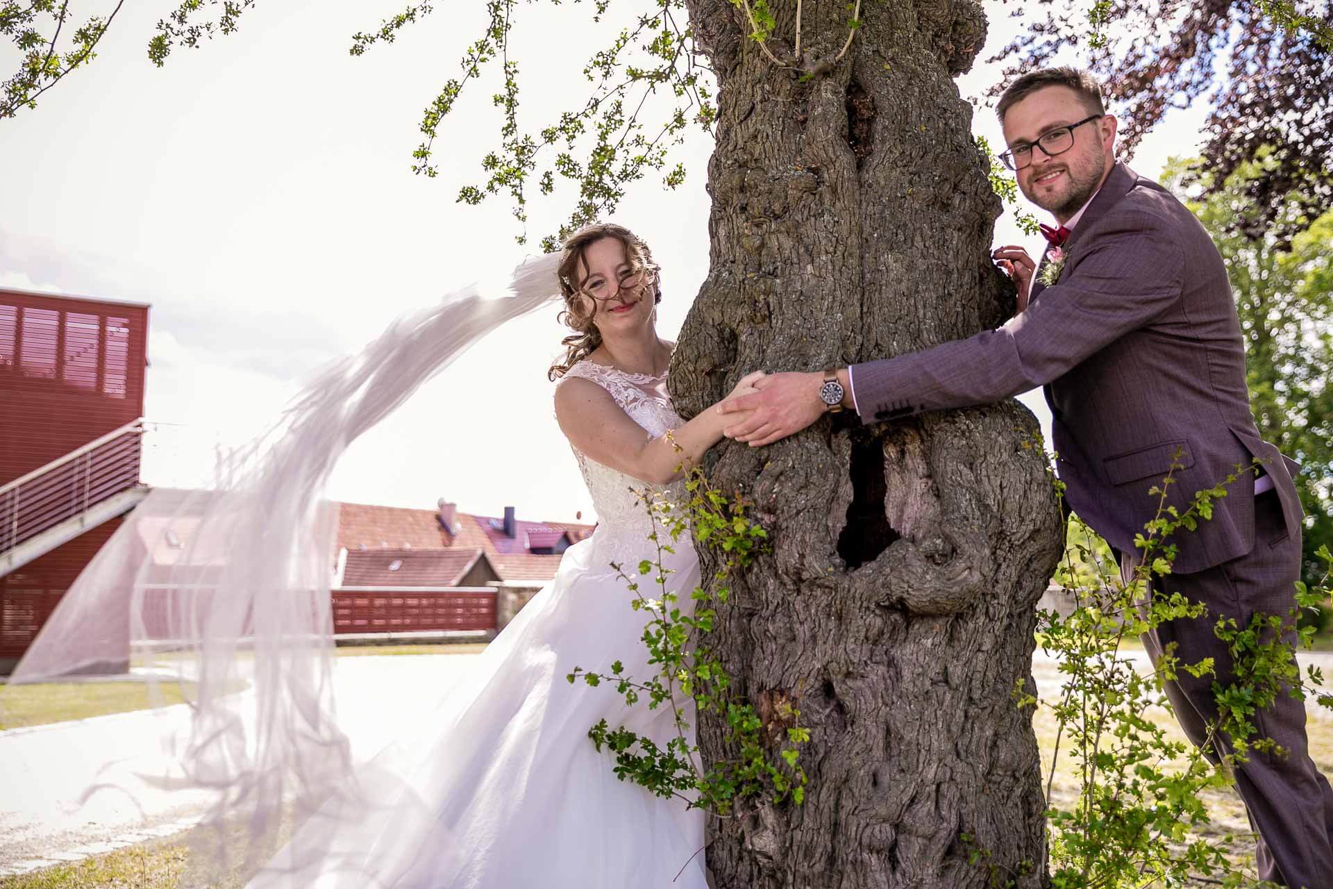 Braut mit fliegenden Schleiher und Bräutigam nach der Trauung schauen hinter einem Baum hervor und halten Händchen